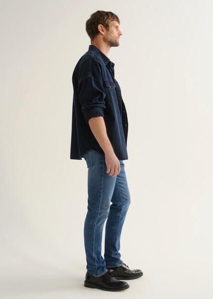 Person wearing a dark navy denim overshirt with rolled sleeves, medium blue slim tapered jeans, and black leather shoes, angled side full body view on a light grey background.