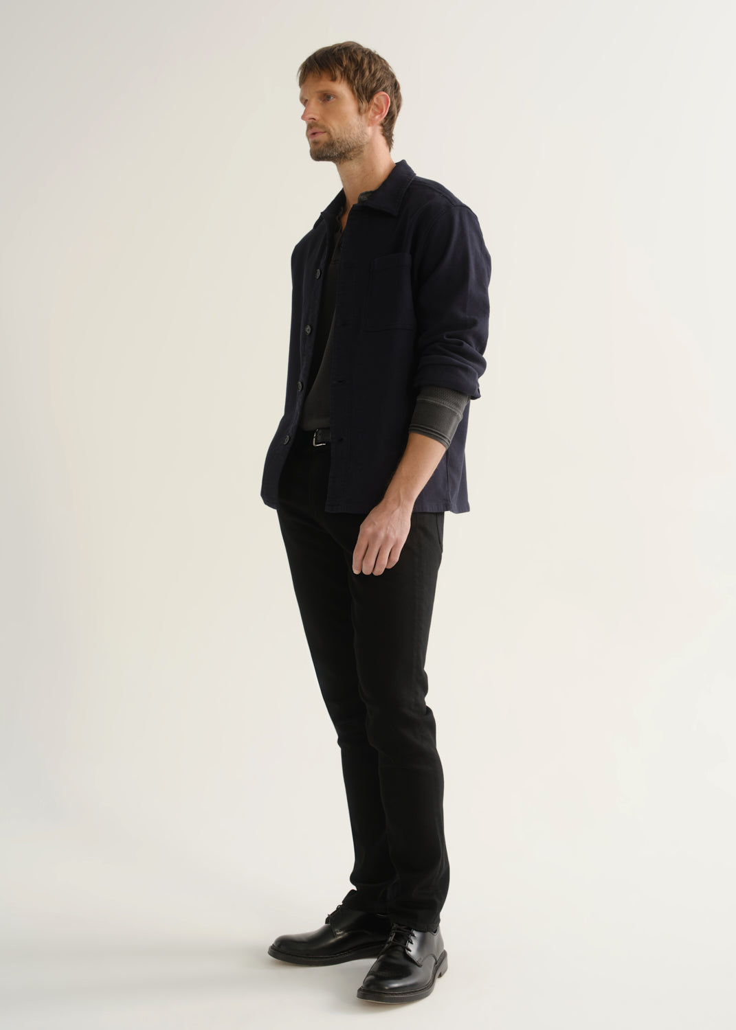 Person wearing black slim tapered jeans with a dark navy button-up overshirt, charcoal grey shirt, and black shoes, front angled view.