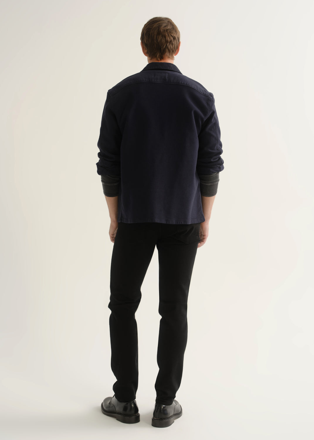 Person wearing black slim tapered jeans with a dark navy button-up overshirt, charcoal grey shirt, and black shoes, back view.