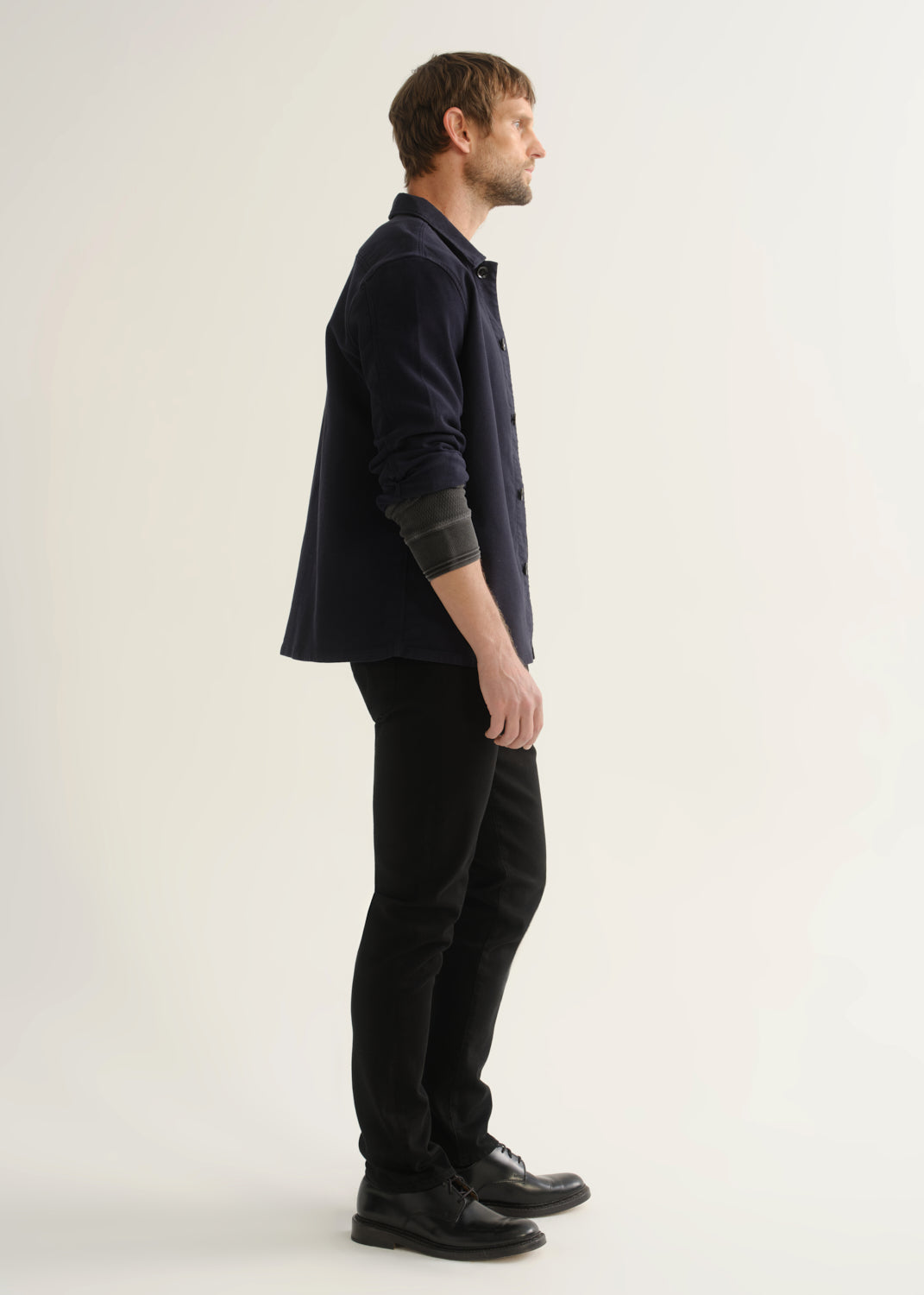 Person wearing black slim tapered jeans with a dark navy button-up overshirt, charcoal grey shirt, and black shoes, side view.