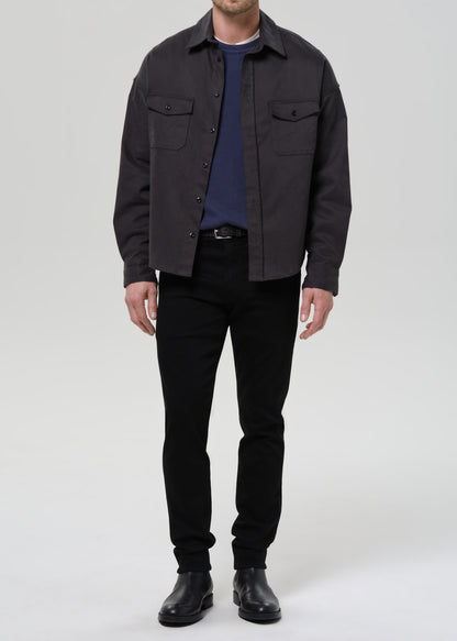 A man wearing a dark charcoal grey overshirt with chest flap pockets, a navy blue t-shirt, black pants, and black leather boots, standing against a grey background.