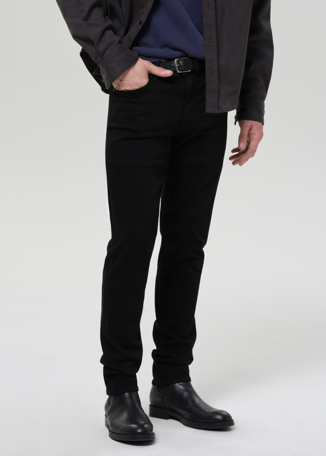 A model wearing slim-fit black jeans, a navy blue shirt, a dark charcoal overshirt, and black leather boots, shown from the waist down against a grey background.