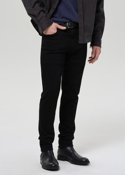 A model wearing slim-fit black jeans, a navy blue shirt, a dark charcoal overshirt, and black leather boots, shown from the waist down against a grey background.