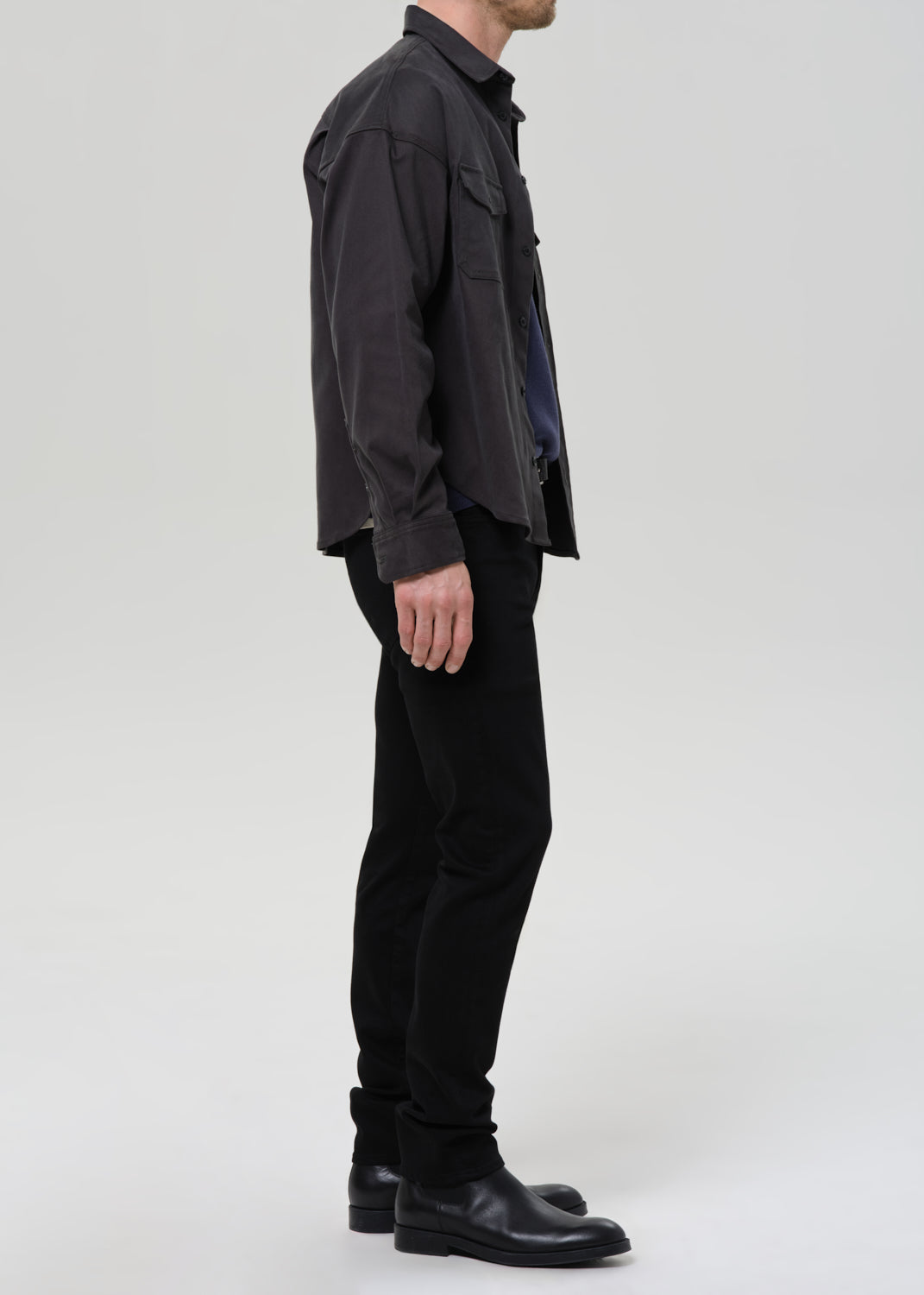 A man shown in profile wearing a dark charcoal gray utility-style overshirt, black trousers, and black leather boots against a grey background.