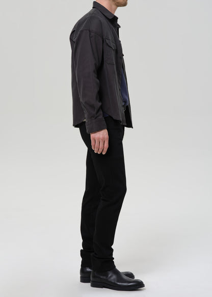 A man shown in profile wearing a dark charcoal gray utility-style overshirt, black trousers, and black leather boots against a grey background.