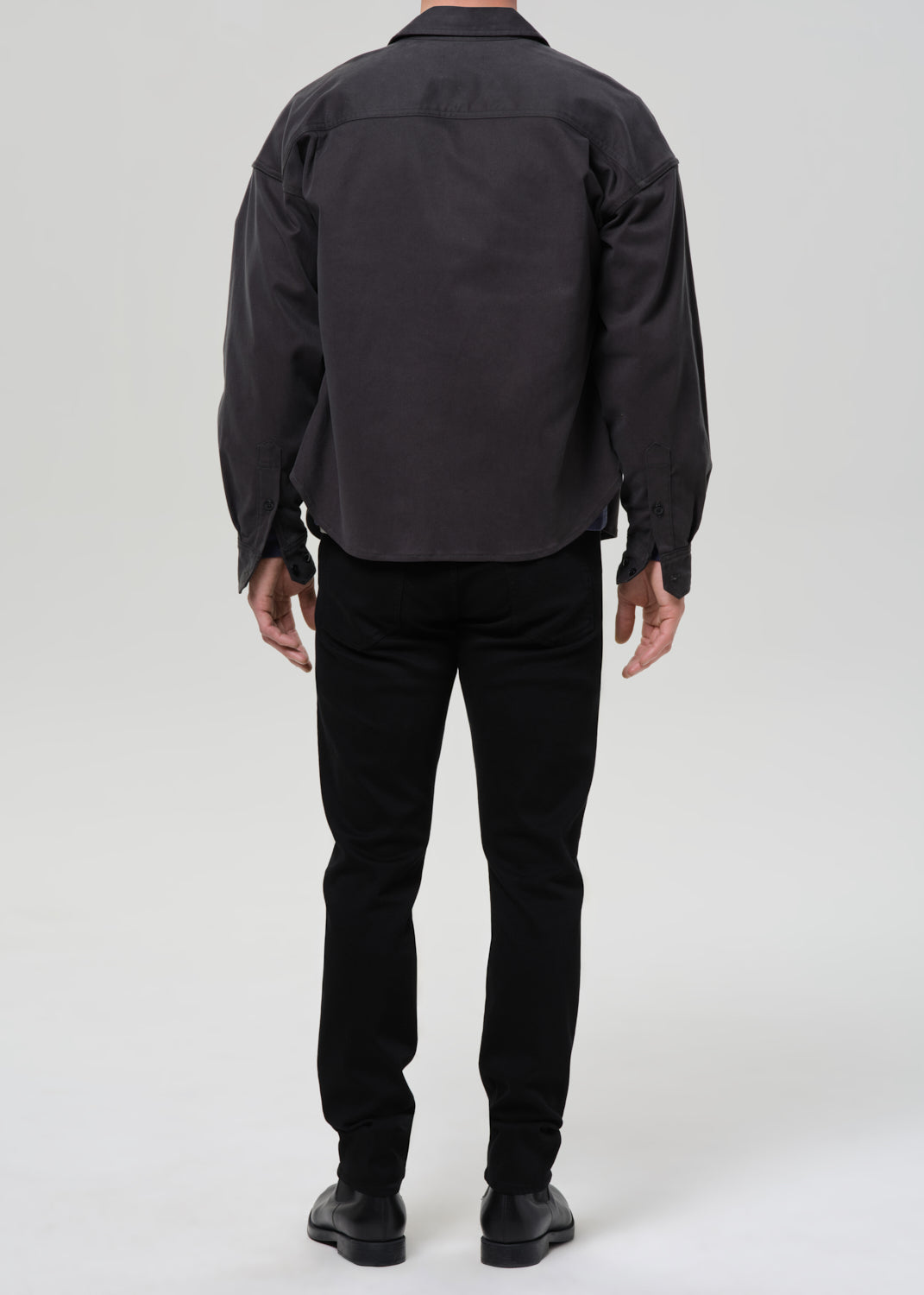 A person shown from the back wearing a dark charcoal gray overshirt, black trousers, and black shoes against a grey background.
