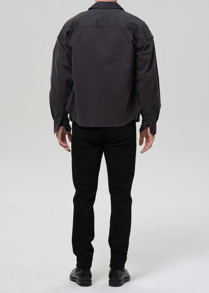 A person shown from the back wearing a dark charcoal gray overshirt, black trousers, and black shoes against a grey background.