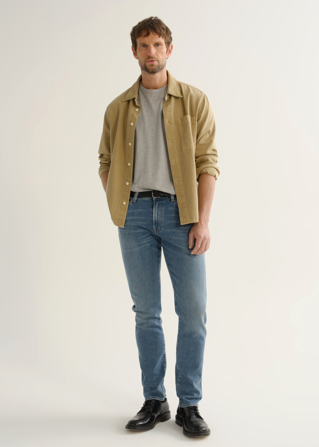 Person wearing a tan button-down overshirt layered over a grey crewneck tee, medium blue slim tapered jeans, and black leather shoes, front full body view on a light grey background.
