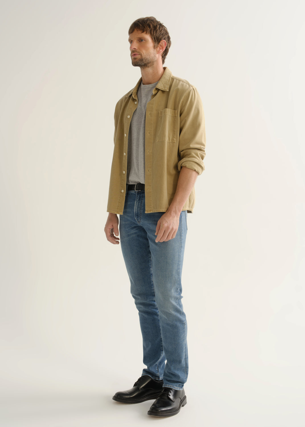 Person wearing a tan button-down overshirt with rolled sleeves layered over a grey crewneck tee, medium blue slim tapered jeans, and black leather shoes, side profile full body view on a light grey background.
