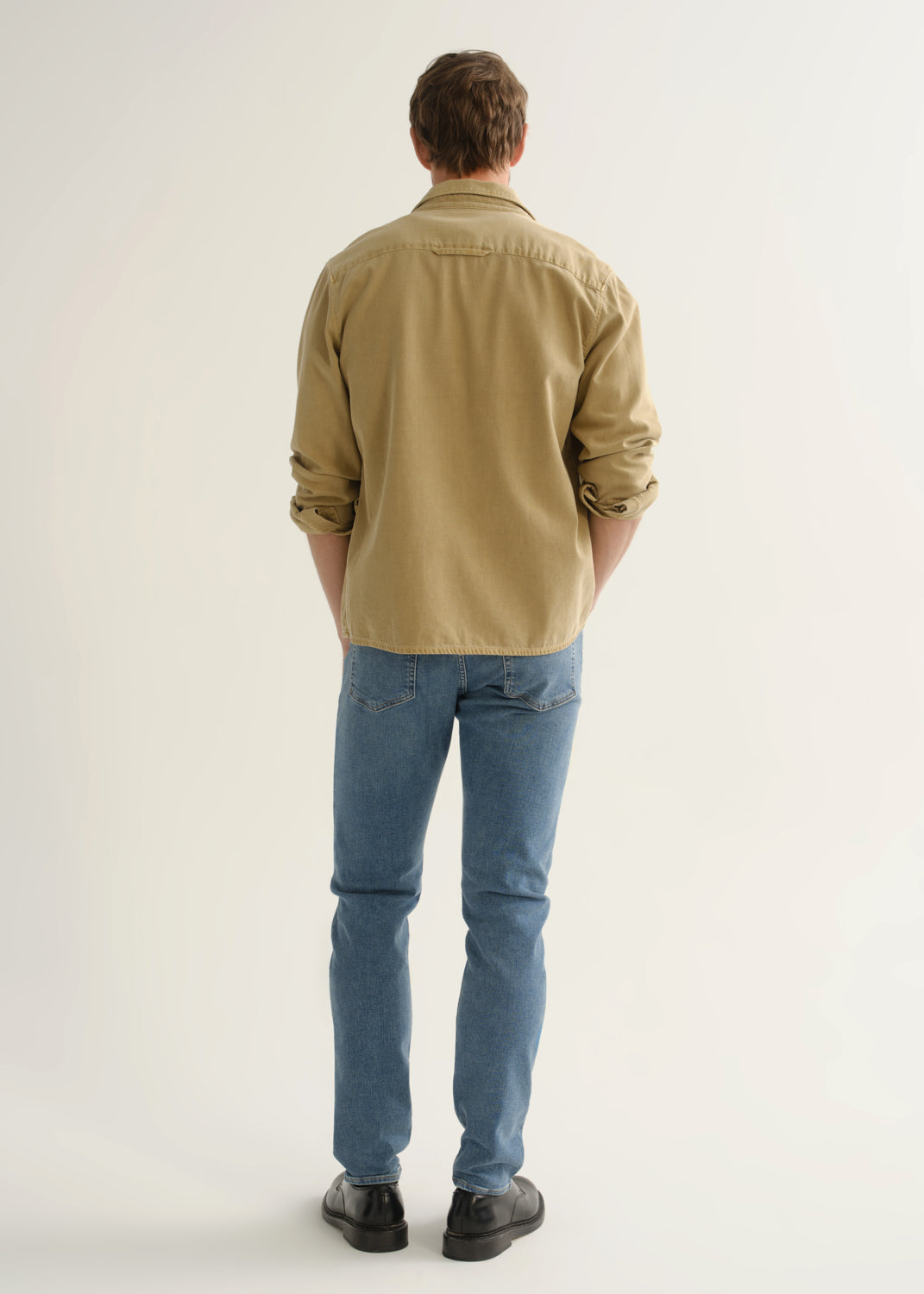 Person wearing a tan button-down overshirt with rolled sleeves, medium blue slim tapered jeans, and black leather shoes, back full body view on a light grey background.