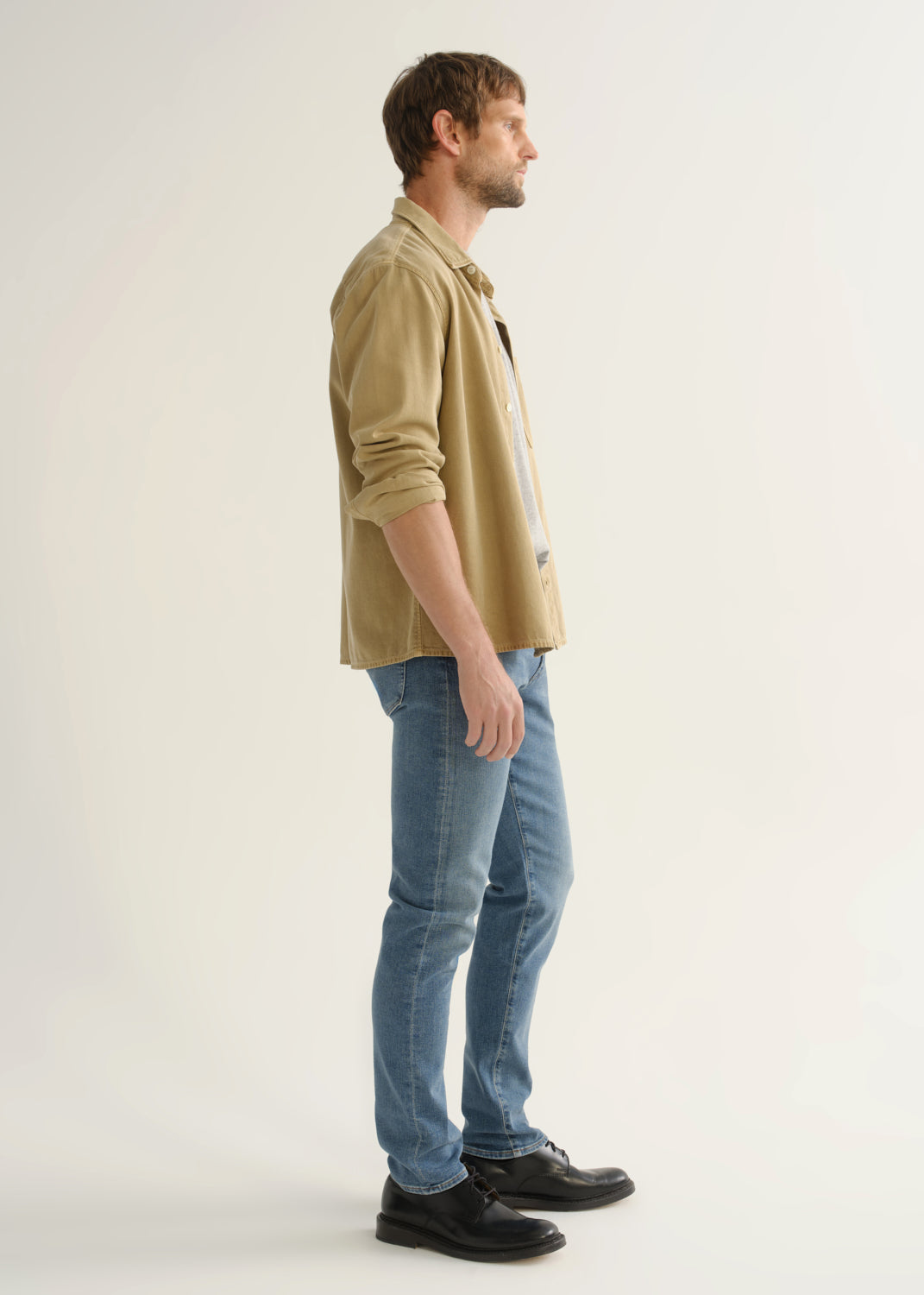 Person wearing a tan button-down overshirt with rolled sleeves, medium blue slim tapered jeans, and black leather shoes, angled side full body view on a light grey background.