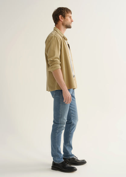 Person wearing a tan button-down overshirt with rolled sleeves, medium blue slim tapered jeans, and black leather shoes, angled side full body view on a light grey background.