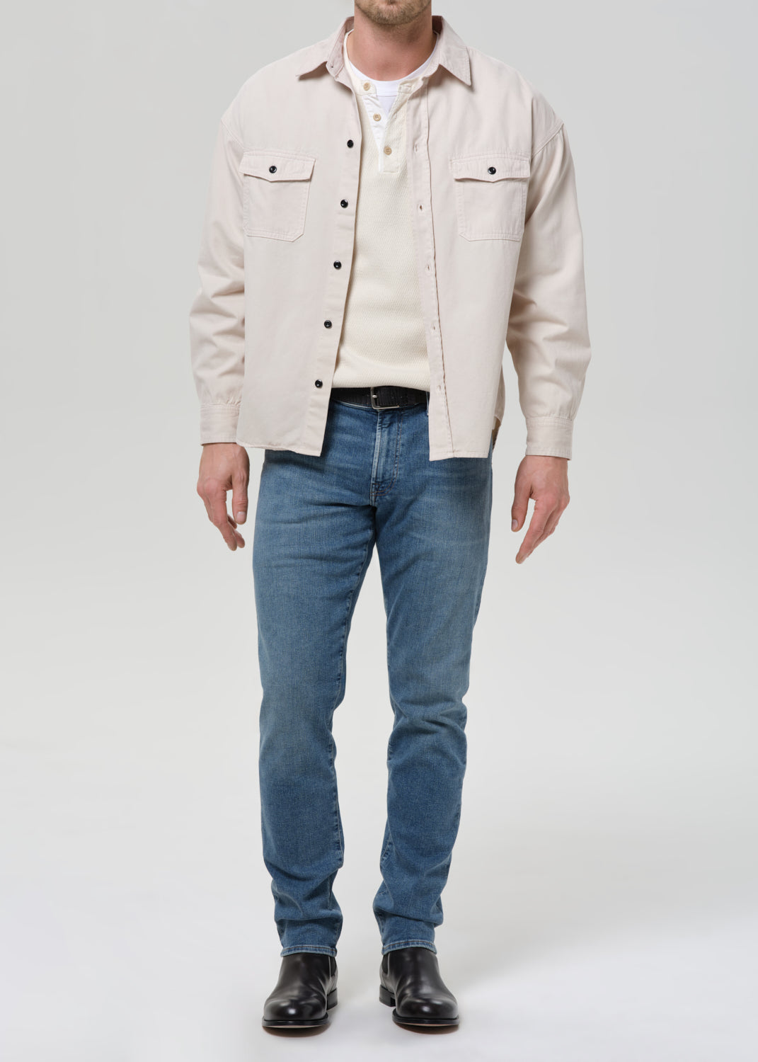A man wearing a light beige overshirt over a cream knit sweater, blue jeans, and black leather boots, standing against a grey background.