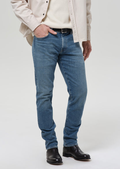 A person wearing medium-wash blue jeans with a cream shirt, a light beige overshirt, and black leather boots, shown from the chest down against a grey background.