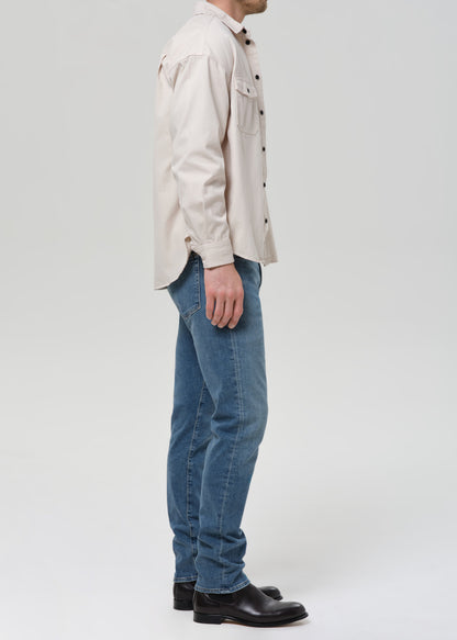 A man shown in profile wearing a light beige button-up shirt, blue jeans, and black leather shoes, photographed against a grey background.