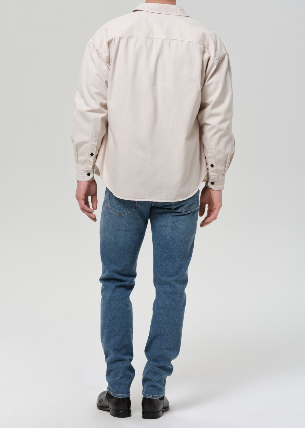 A man viewed from behind wearing a light beige long-sleeve button-up shirt and blue jeans, standing against a grey background.