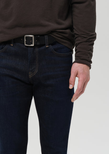 Close-up of a person wearing dark blue slim-fit jeans with a black belt and silver buckle, paired with a dark brown long-sleeve top, against a grey background.