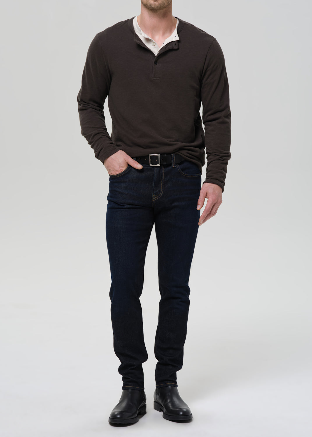 A man viewed from the front wearing a dark brown long-sleeve shirt, dark blue jeans, and black shoes, standing against a grey background.