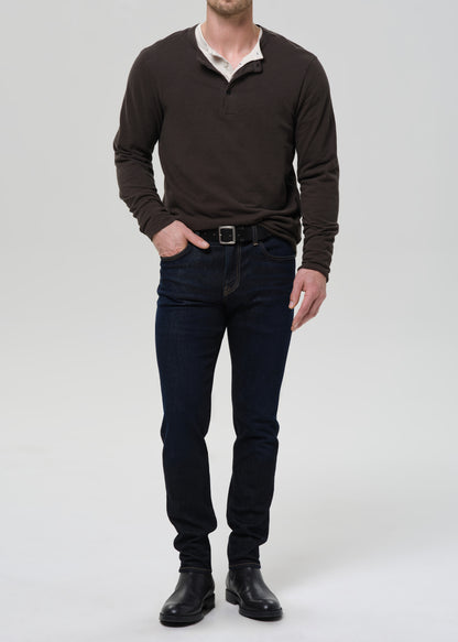 A man viewed from the front wearing a dark brown long-sleeve shirt, dark blue jeans, and black shoes, standing against a grey background.