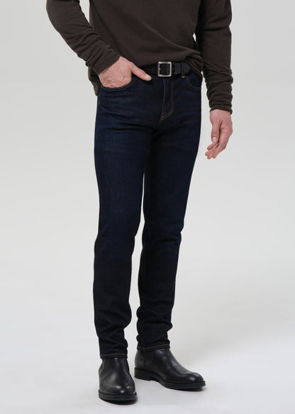 A model wearing dark navy slim-fit jeans, a dark brown long-sleeve shirt, a black belt, and black leather boots, standing against a grey background.