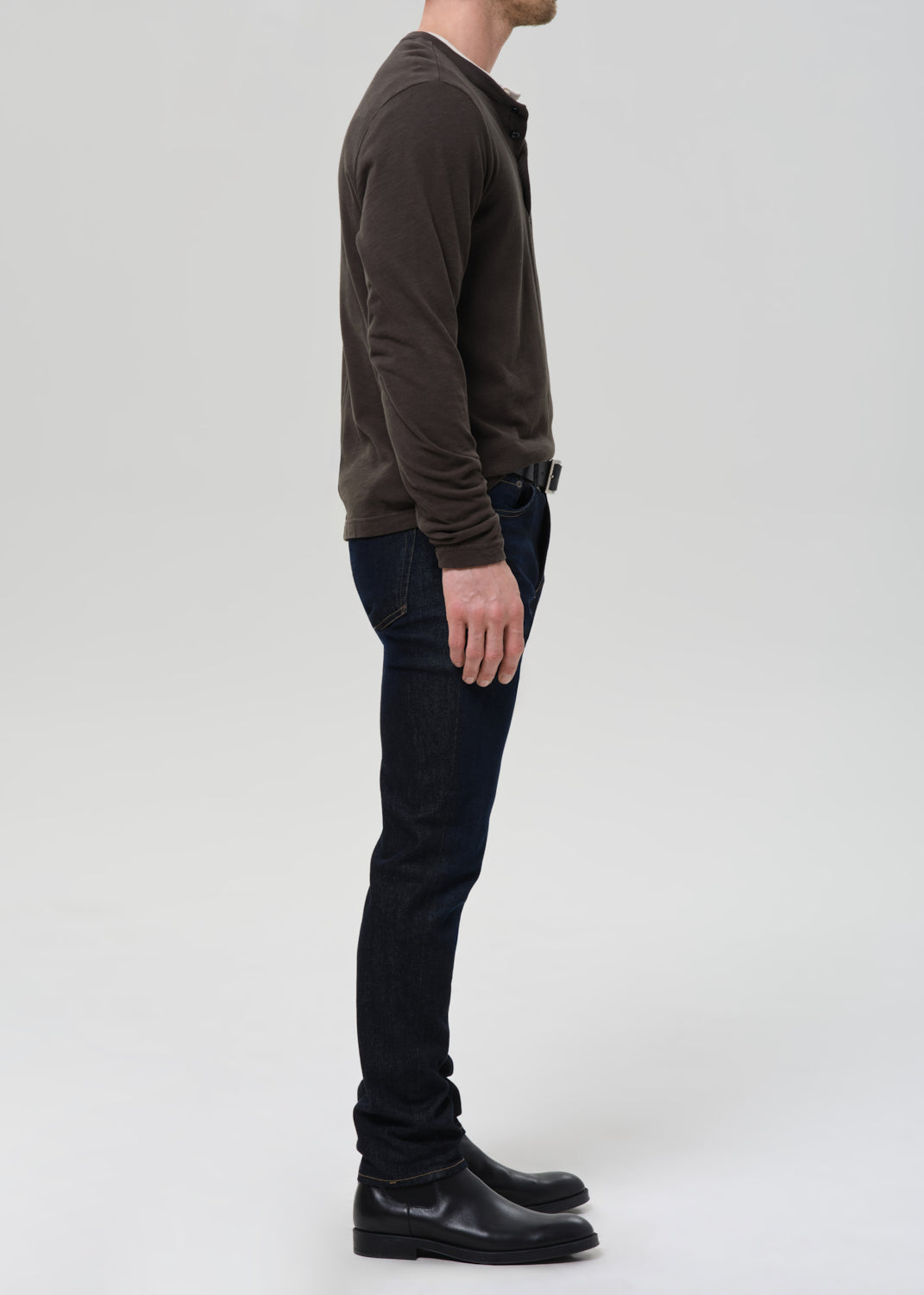 A man shown in profile wearing a dark brown long-sleeve knit top, dark blue jeans, and black leather boots against a grey background.