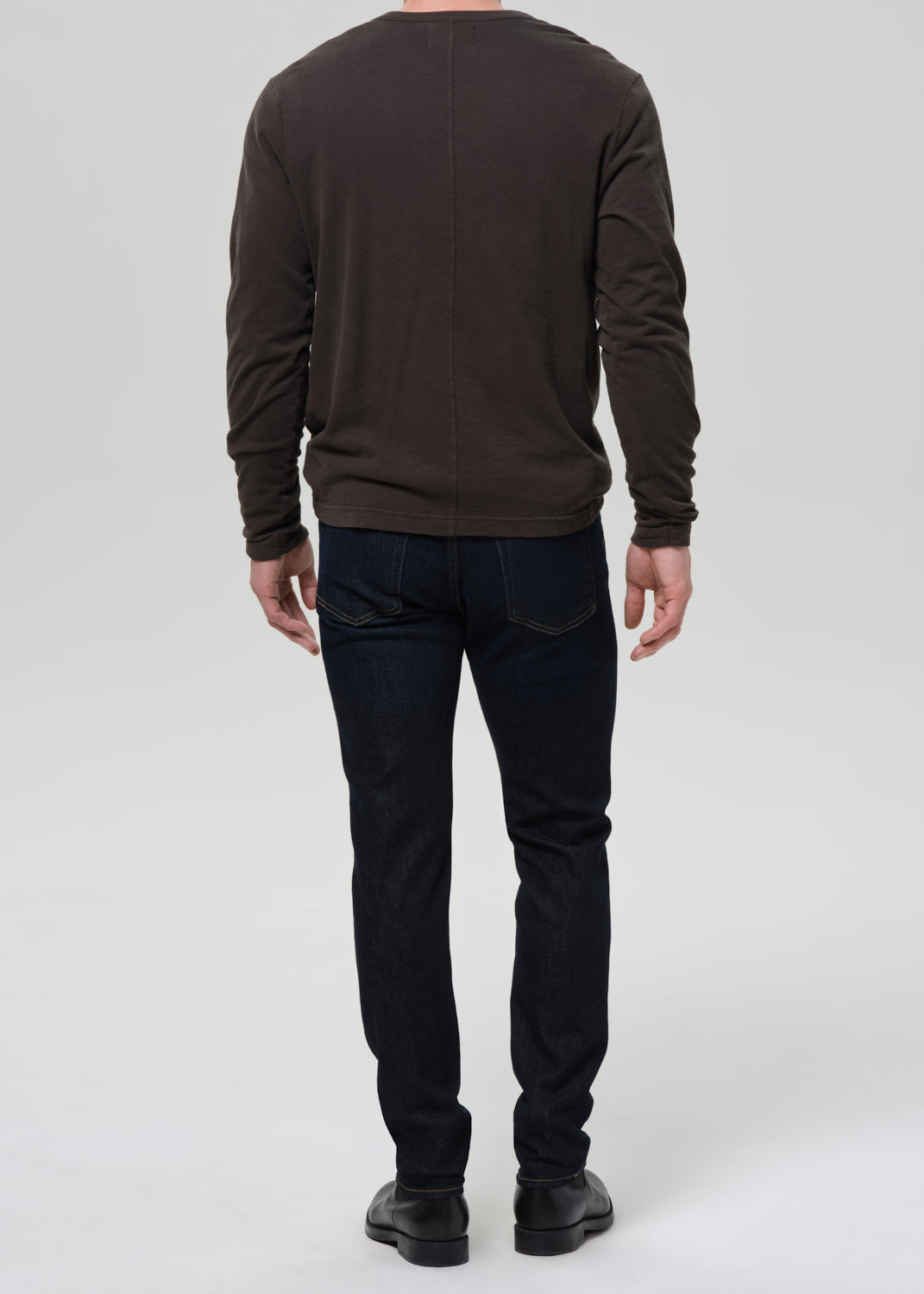 A man viewed from the back wearing a dark brown long-sleeve shirt, dark blue jeans, and black shoes, standing against a grey background.