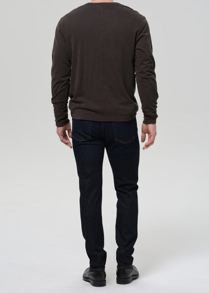A man viewed from the back wearing a dark brown long-sleeve shirt, dark blue jeans, and black shoes, standing against a grey background.