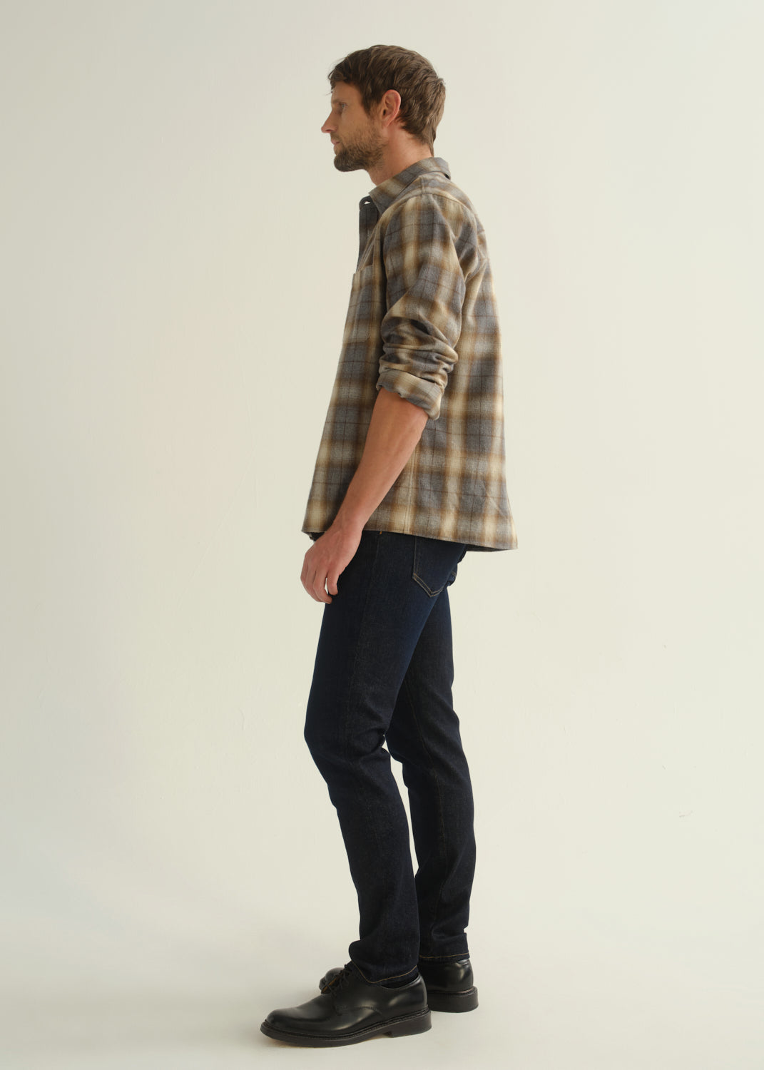 Person wearing a grey and brown plaid button-down shirt with rolled sleeves layered over a black crewneck tee, dark indigo slim tapered jeans, and black leather shoes, angled front full body view on a light grey background.