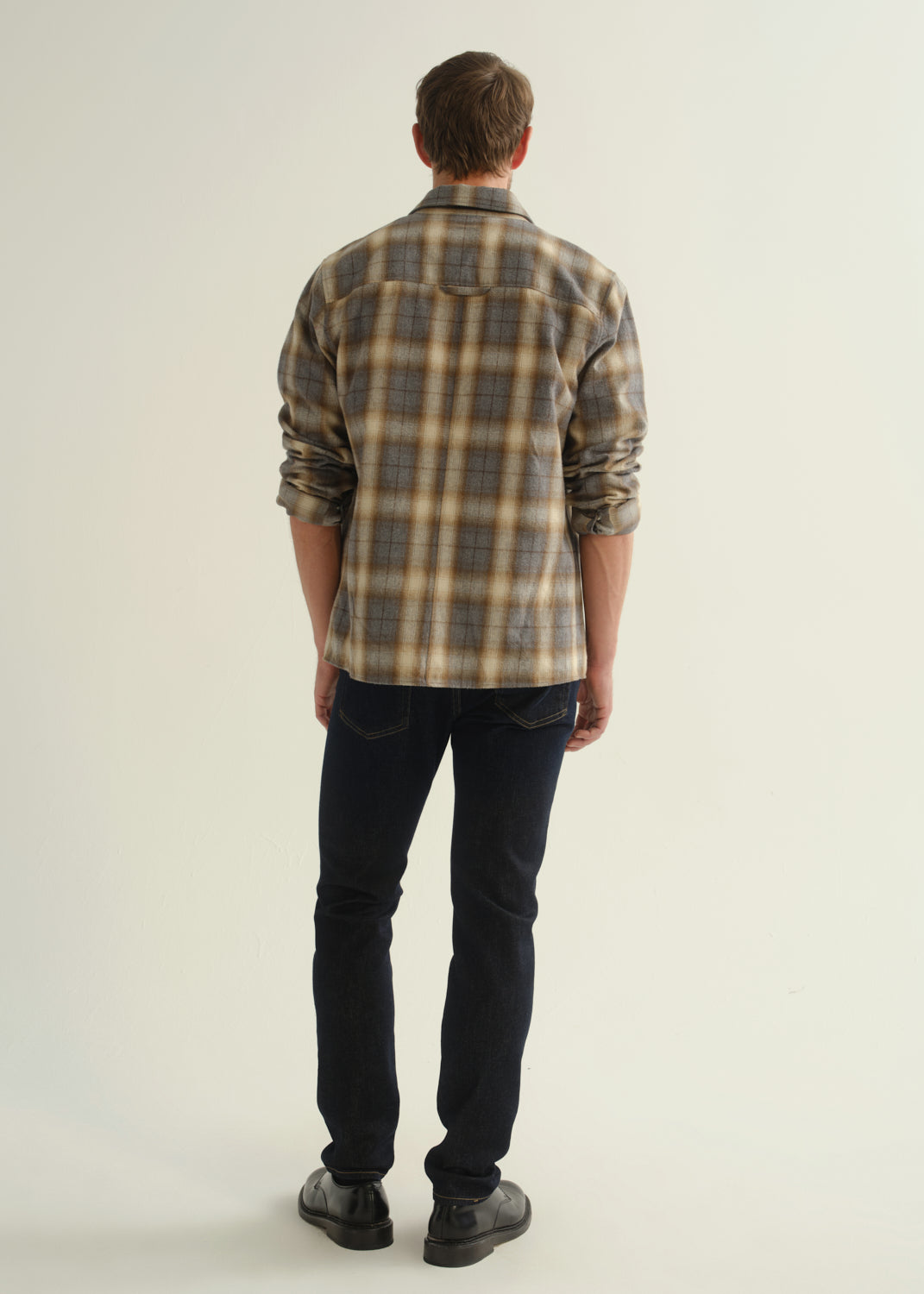 Person wearing a grey and brown plaid button-down shirt with rolled sleeves, dark indigo slim tapered jeans, and black leather shoes, back full body view on a light grey background.
