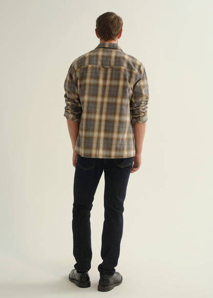Person wearing a grey and brown plaid button-down shirt with rolled sleeves, dark indigo slim tapered jeans, and black leather shoes, back full body view on a light grey background.