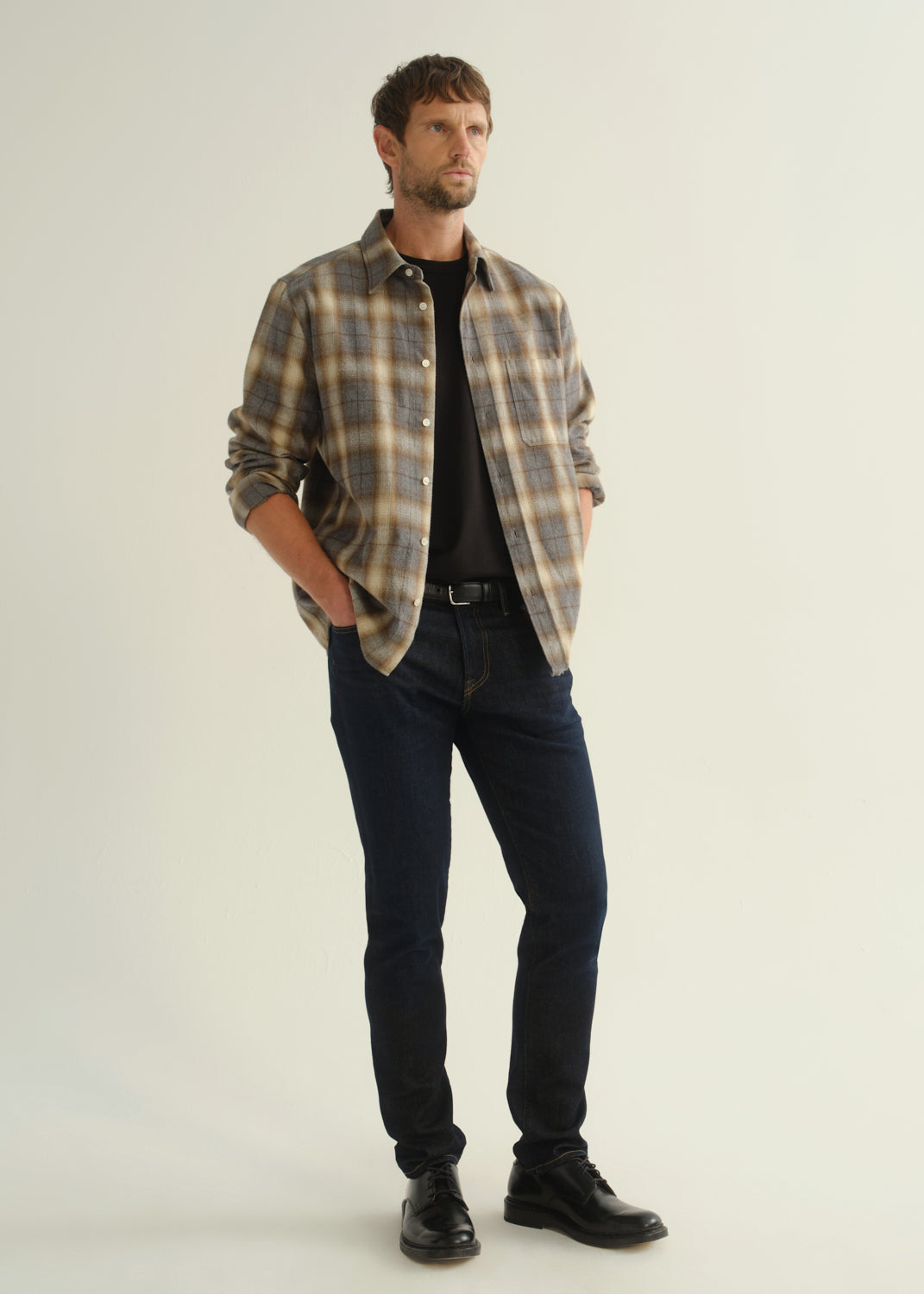 Person wearing a grey and brown plaid button-down shirt with rolled sleeves, dark indigo slim tapered jeans, and black leather shoes, side full body view on a light grey background.