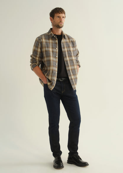 Person wearing a grey and brown plaid button-down shirt with rolled sleeves, dark indigo slim tapered jeans, and black leather shoes, side full body view on a light grey background.