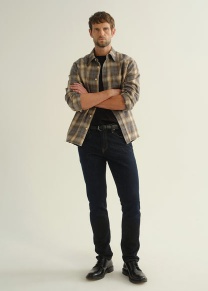 Person wearing a grey and brown plaid button-down shirt with rolled sleeves layered over a black crewneck tee, dark indigo slim tapered jeans, and black leather shoes, front full body view on a light grey background.