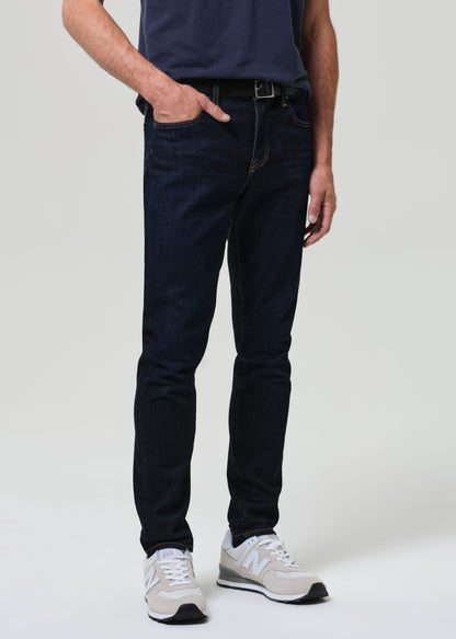 Man wearing a navy t-shirt and dark wash jeans against a grey background