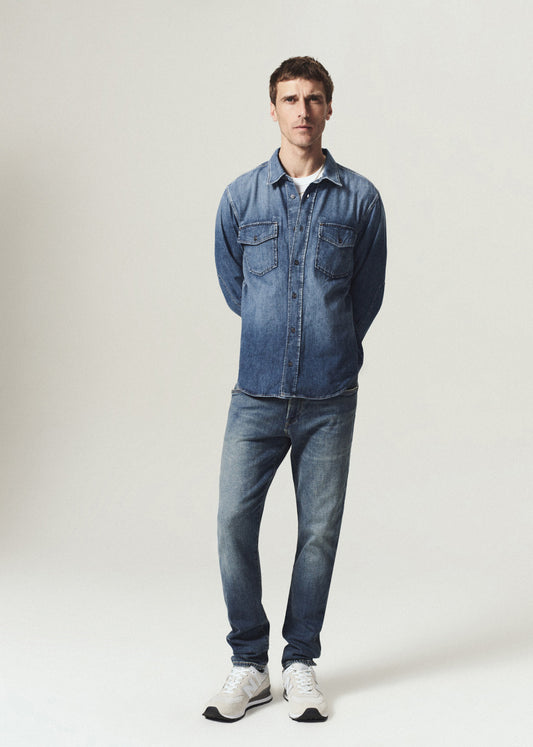 A man modeling a blue denim button-up shirt and matching blue jeans with white sneakers, standing against a light gray studio background.