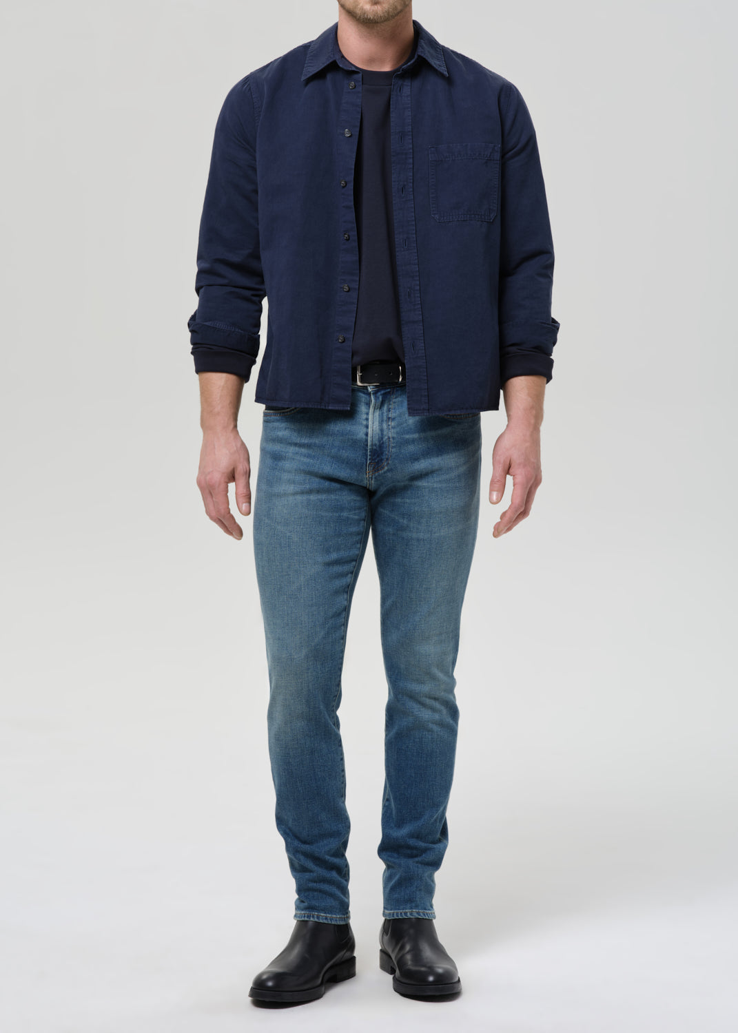 A man wearing a dark navy blue button-up shirt over a black t-shirt, blue jeans, and black leather boots, standing against a grey background.