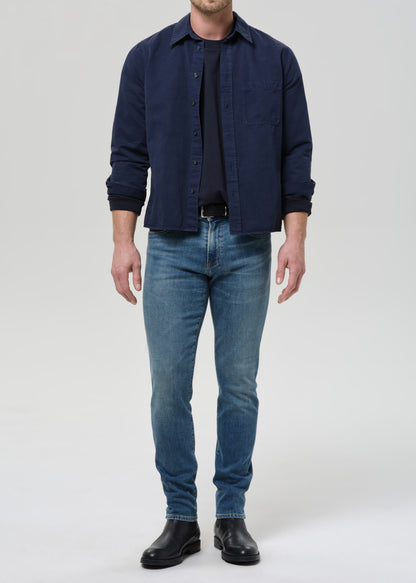 A man wearing a dark navy blue button-up shirt over a black t-shirt, blue jeans, and black leather boots, standing against a grey background.
