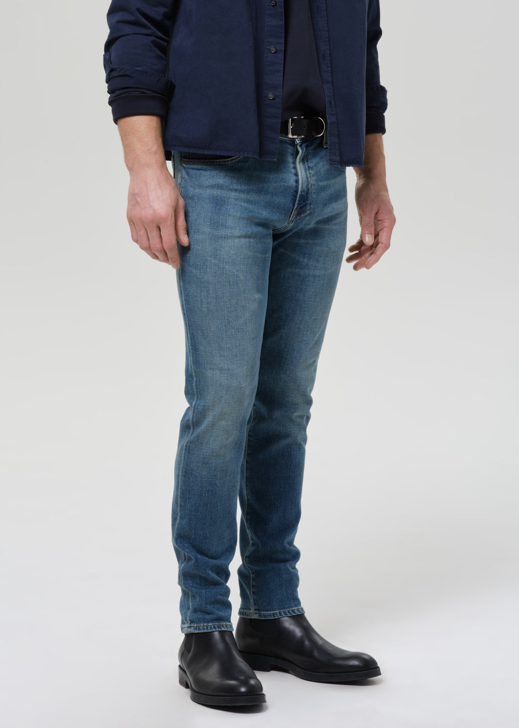 A man wearing slim-fit medium wash blue jeans, a navy blue button-up shirt, and black leather boots, standing against a grey background.