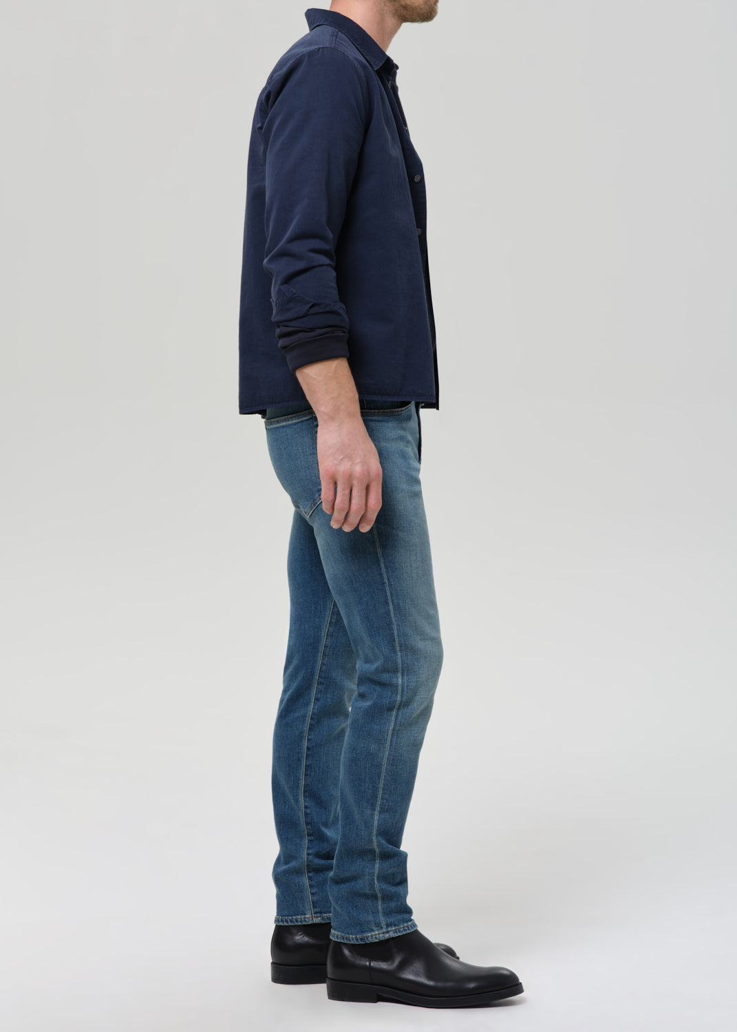 A man viewed from the side wearing a navy blue jacket, blue jeans, and black shoes, standing against a grey background.