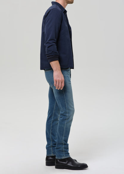 A man viewed from the side wearing a navy blue jacket, blue jeans, and black shoes, standing against a grey background.