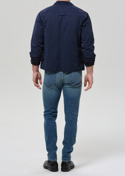 A man viewed from the back wearing a navy blue jacket, blue jeans, and black shoes, standing against a grey background.