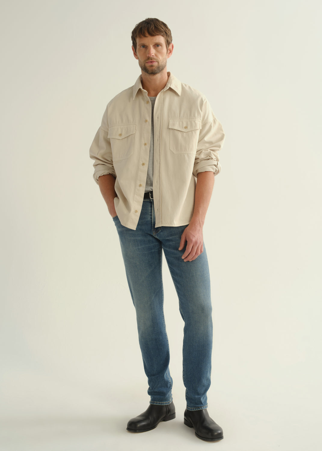 Person wearing a cream button-down shirt with flap chest pockets and rolled sleeves layered over a grey crewneck tee, medium blue slim tapered jeans, and black leather boots, angled front full body view on a light grey background.