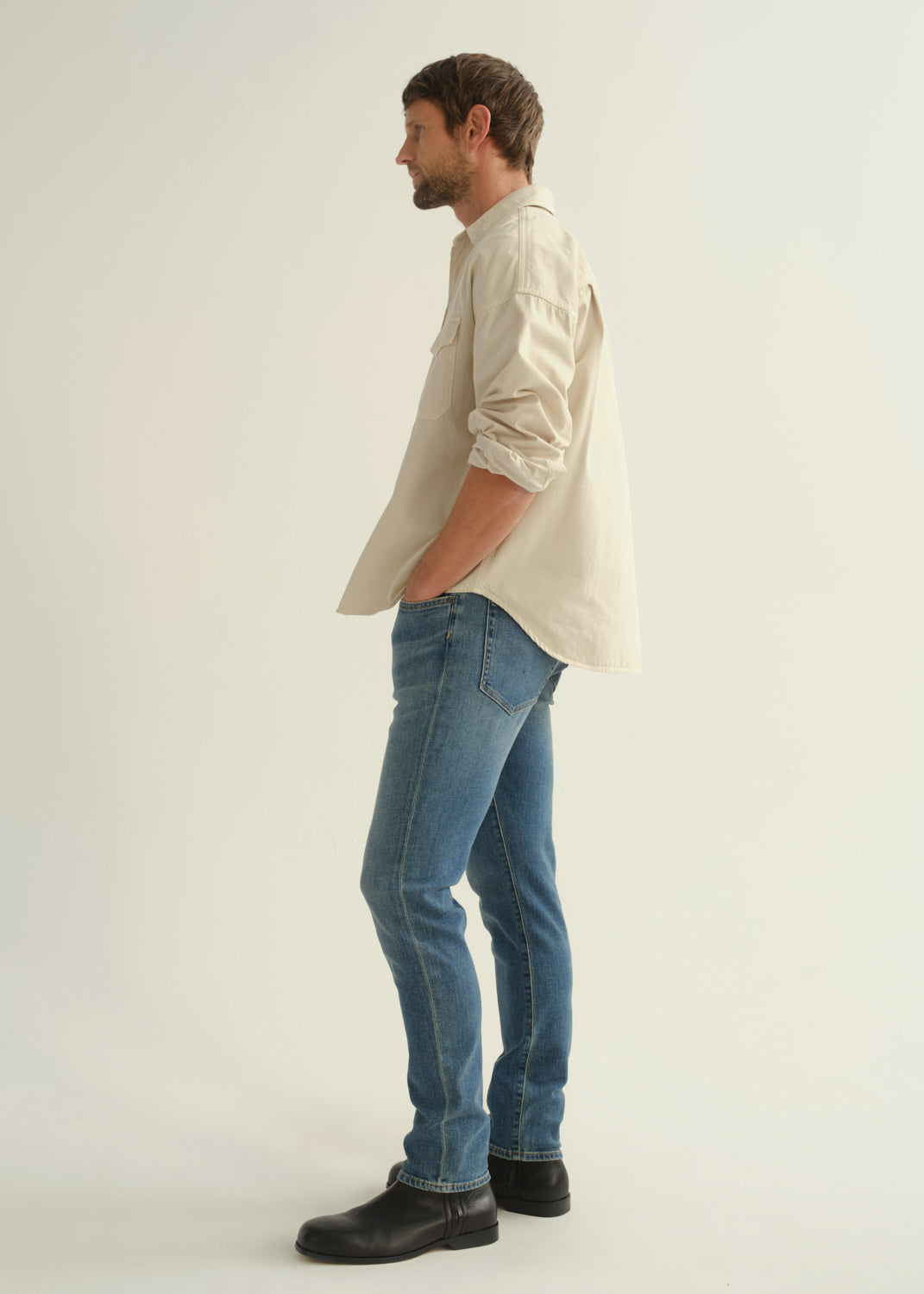 Person wearing a cream button-down shirt with rolled sleeves, medium blue slim tapered jeans, and black leather boots, side full body view on a light grey background.