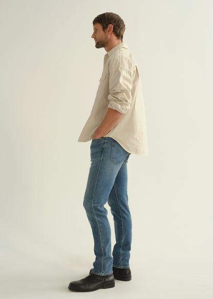Person wearing a cream button-down shirt with rolled sleeves, medium blue slim tapered jeans, and black leather boots, side full body view on a light grey background.