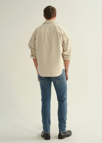 Person wearing a cream button-down shirt with rolled sleeves, medium blue slim tapered jeans, and black leather boots, back full body view on a light grey background.
