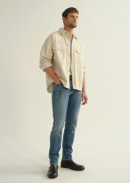 Person wearing a cream button-down shirt with flap chest pockets and rolled sleeves layered over a grey crewneck tee, medium blue slim tapered jeans, and black leather boots, front full body view on a light grey background.