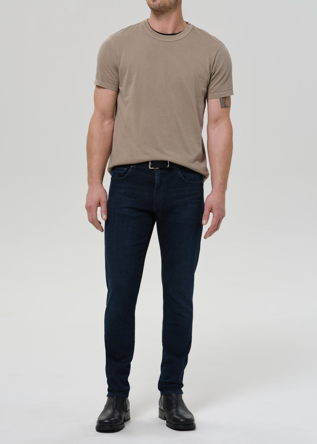 Man wearing a beige t-shirt and dark jeans on a white background