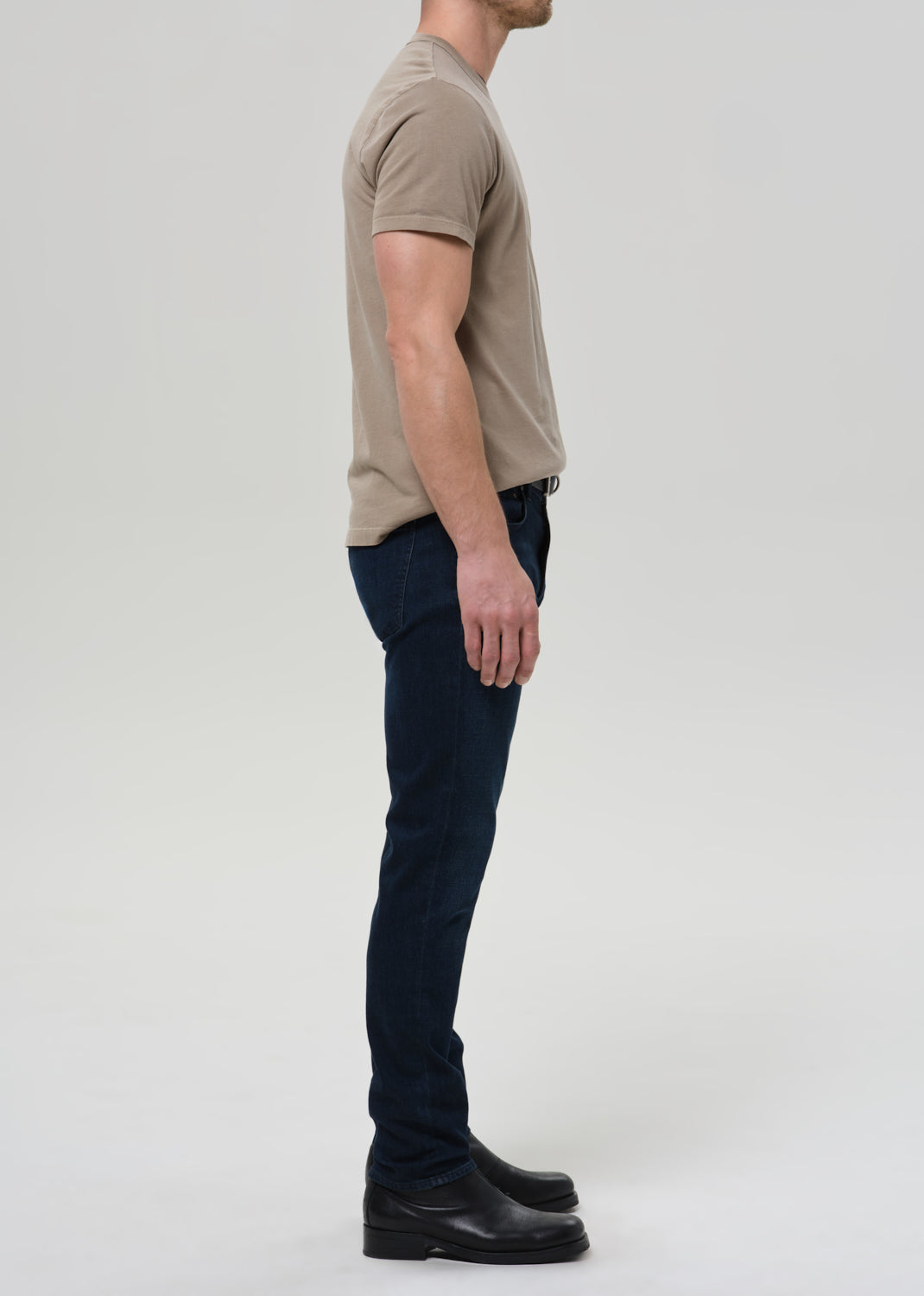 Man wearing a beige t-shirt and dark jeans standing against a plain background