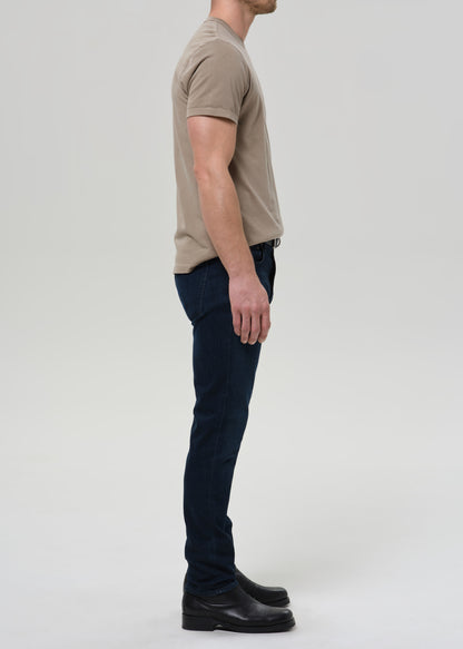 Man wearing a beige t-shirt and dark jeans standing against a plain background