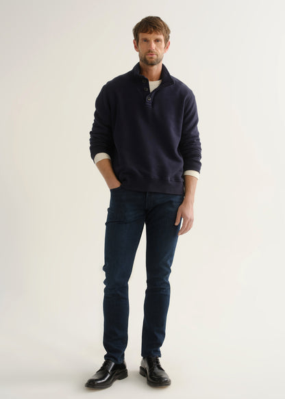 Person wearing a navy long-sleeve pullover with a mock collar and button placket layered over a white tee, dark indigo slim tapered jeans, and black leather shoes, angled front full body view on a light grey background.